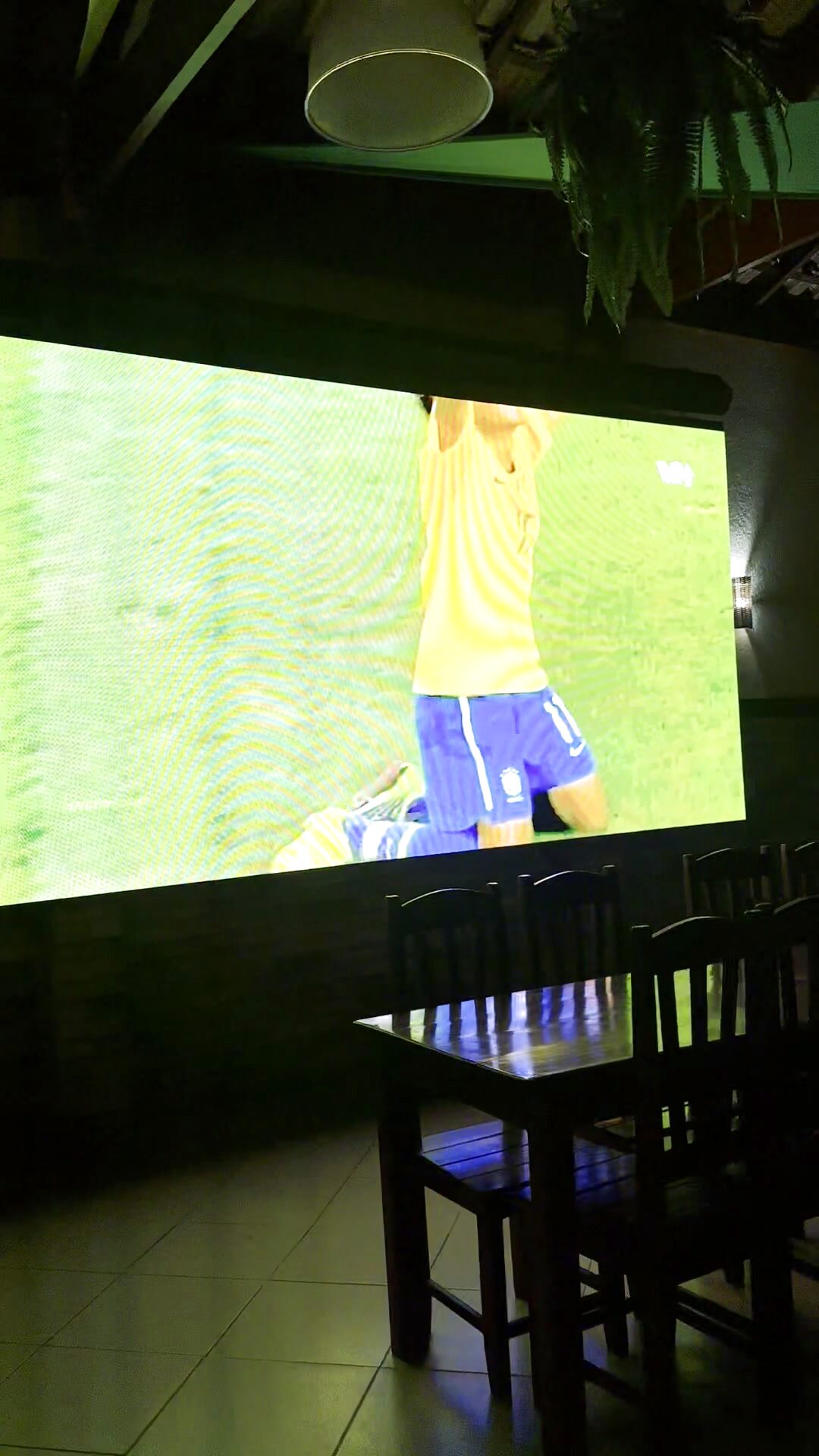 Salão da Kabana com telão exibindo um jogo e mesas de madeira